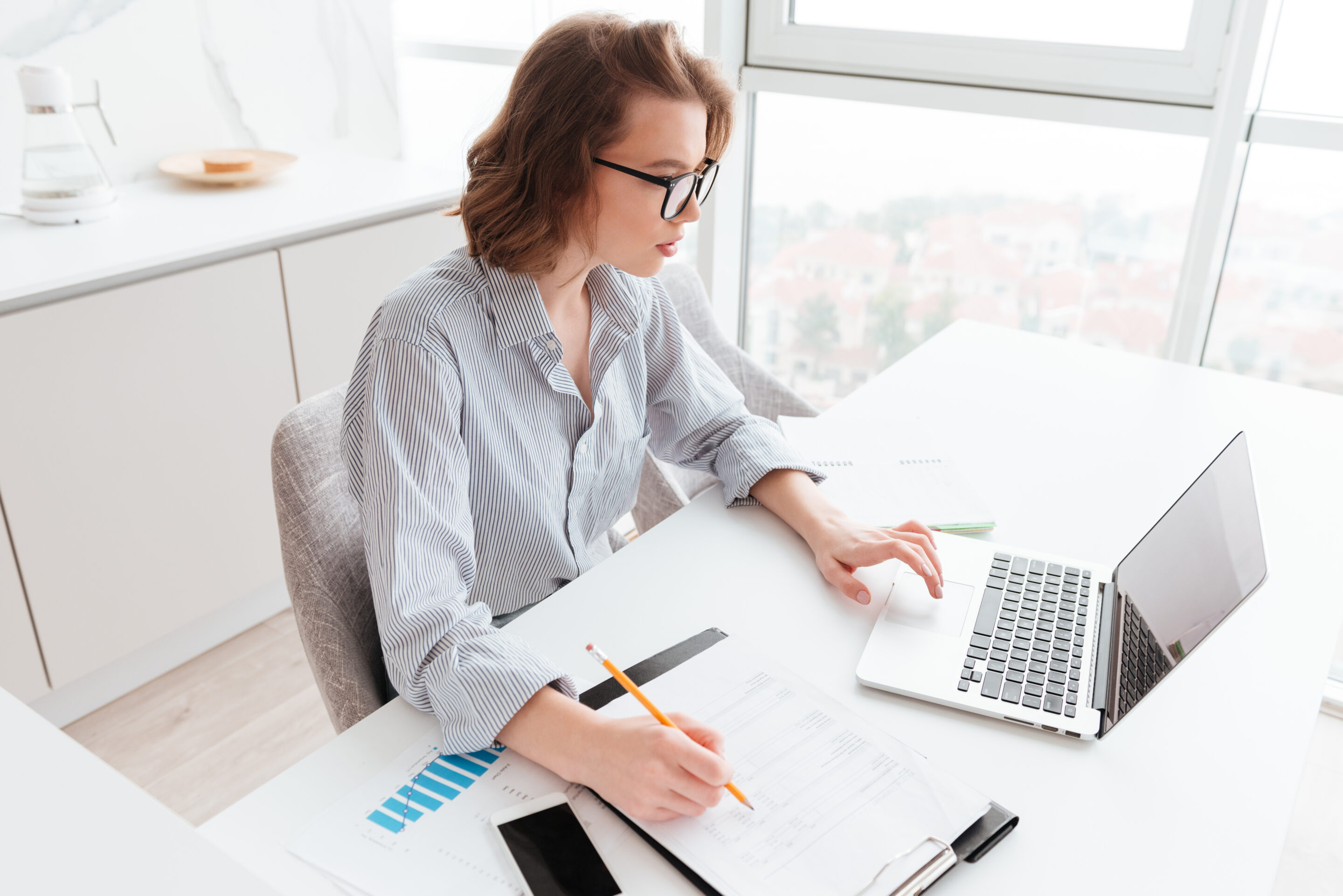 charming caucasian businesswoman striped shirt working with laptop papers light apartment